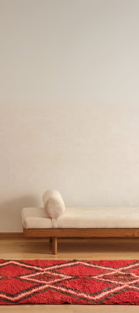 A minimalist room showcasing a wooden bench with a cream cushion and cylindrical pillow, set against a plain wall. A vintage kilim rug, with its intricate patterns, partially appears in the foreground.